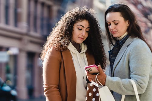 Young girlfriends in warm coats and gloves standing in street with gift packages while looking at information on cellphone near buildings in town in sunny day